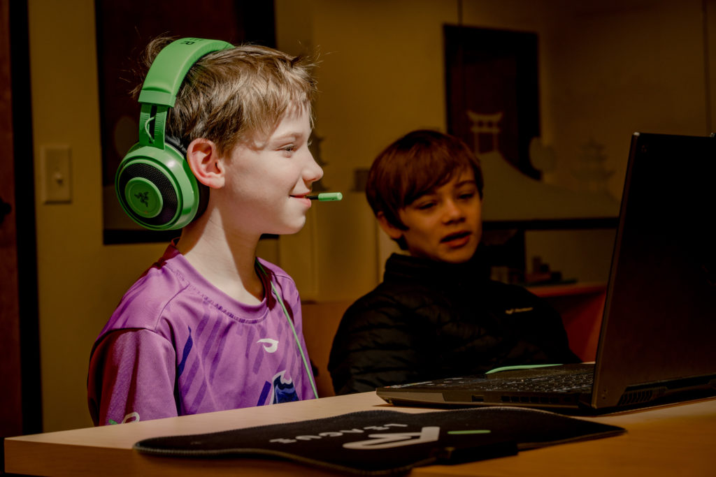 Brysyn Monck sonríe durante un juego de Fortnite, mirando la pantalla de su computadora portátil mientras Kayden Seeley se sienta a su izquierda, colaborando en la acción.