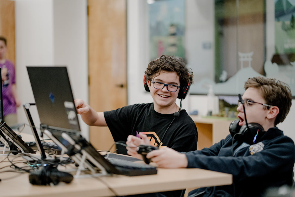 Dos adolescentes interactúan, uno sonriendo y el otro expresando sorpresa, mientras juegan Fortnite en una reunión de la Liga XP.