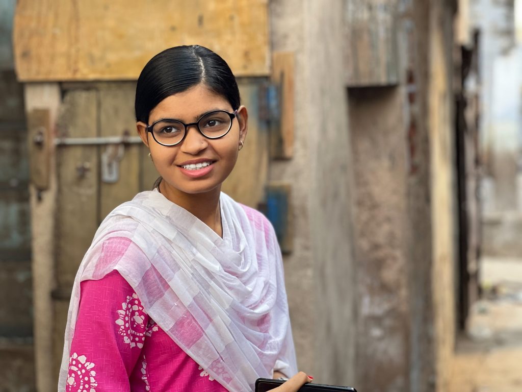 Una niña con anteojos en un pueblo sonríe lejos de la cámara.