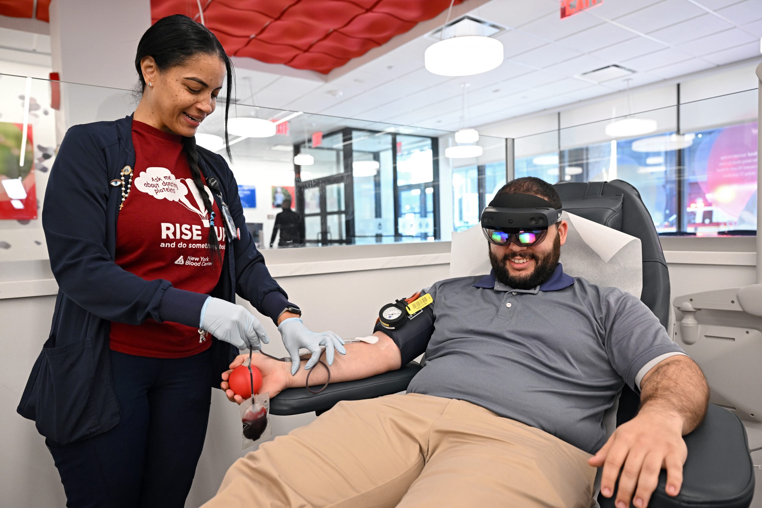 Un miembro del personal en un sitio de donación de sangre recolecta sangre de un donante sentado que usa una diadema HoloLens 2 de realidad mixta.