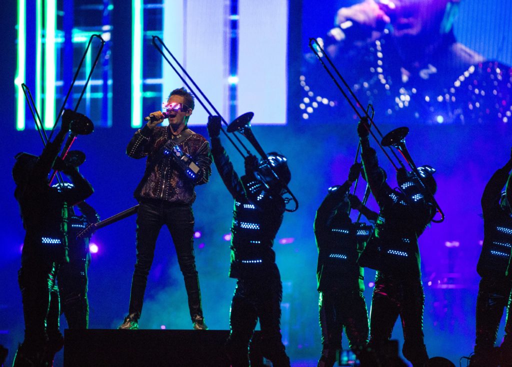 Vocalista da banda Muse canta no palco com óculos de realidade virtual.
