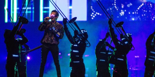 Vocalista da banda Muse canta no palco com óculos de realidade virtual.