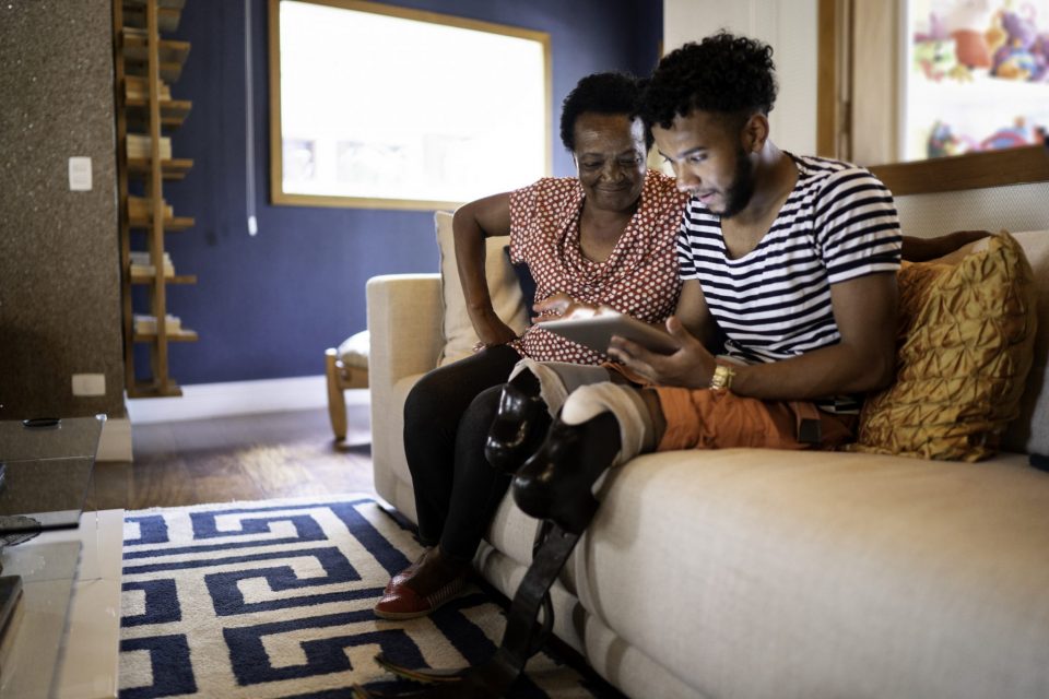 Um jovem com deficiência física utiliza um tablet junto com sua avó, sentados em um sofá em uma casa no Brasil