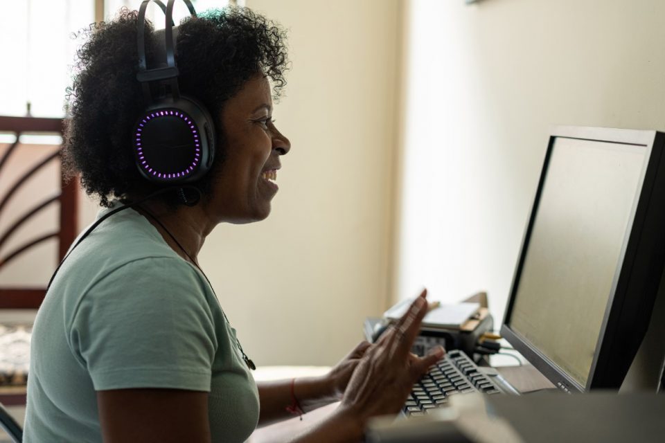 mulher de perfil, usa fones de ouvido enquanto olha para a rela de um computador e sorri