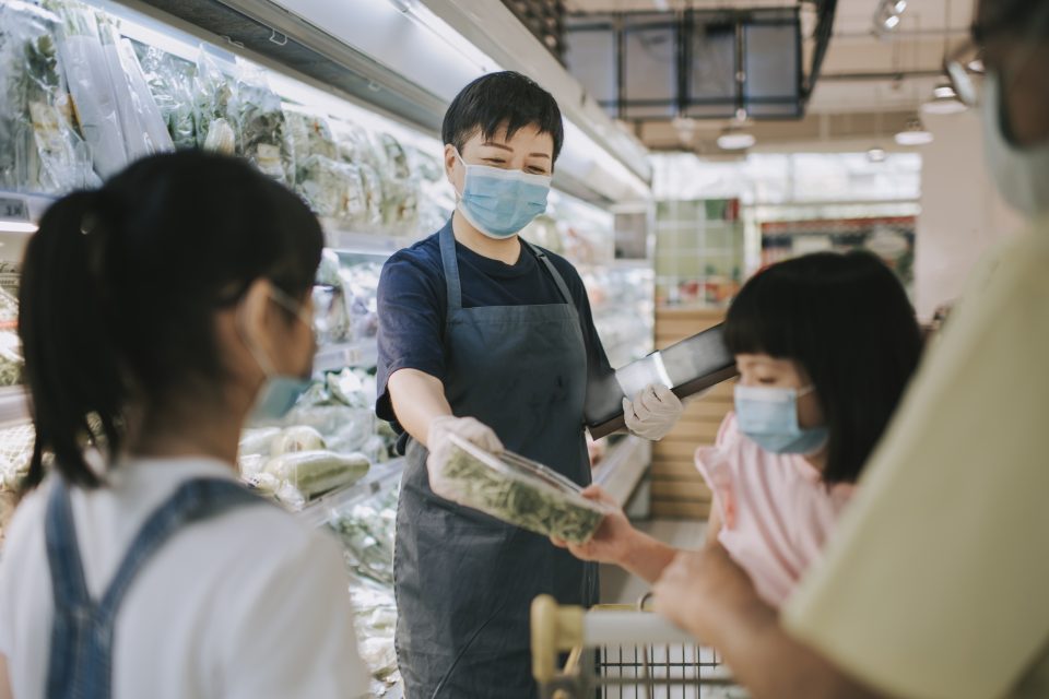 An Asian Chinese female retail assistant advising and helping a family to select the vegetable from the refrigerated section in supermarket. All are wearing masks.