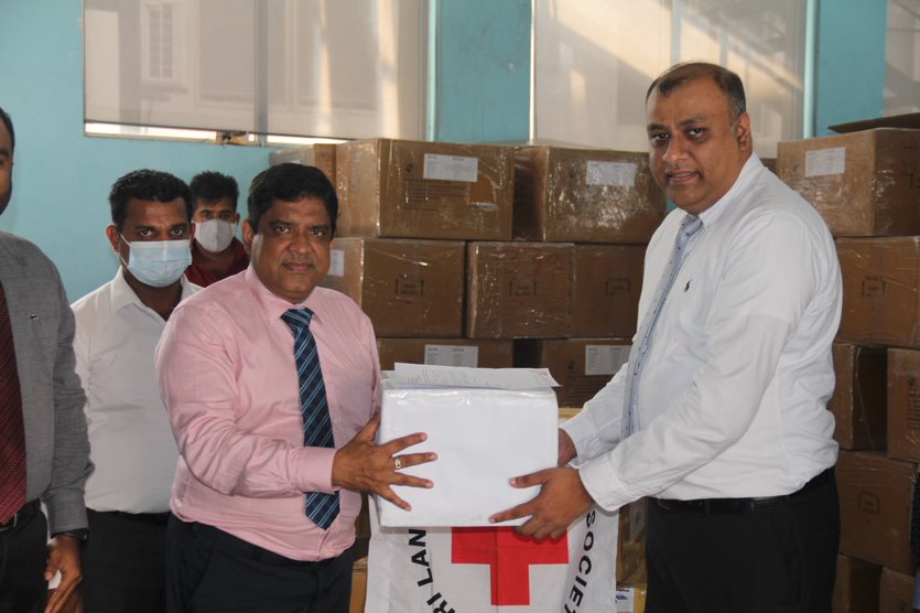 Two men holding a box with the red cross flag and boxes of medical supplies in the backdrop