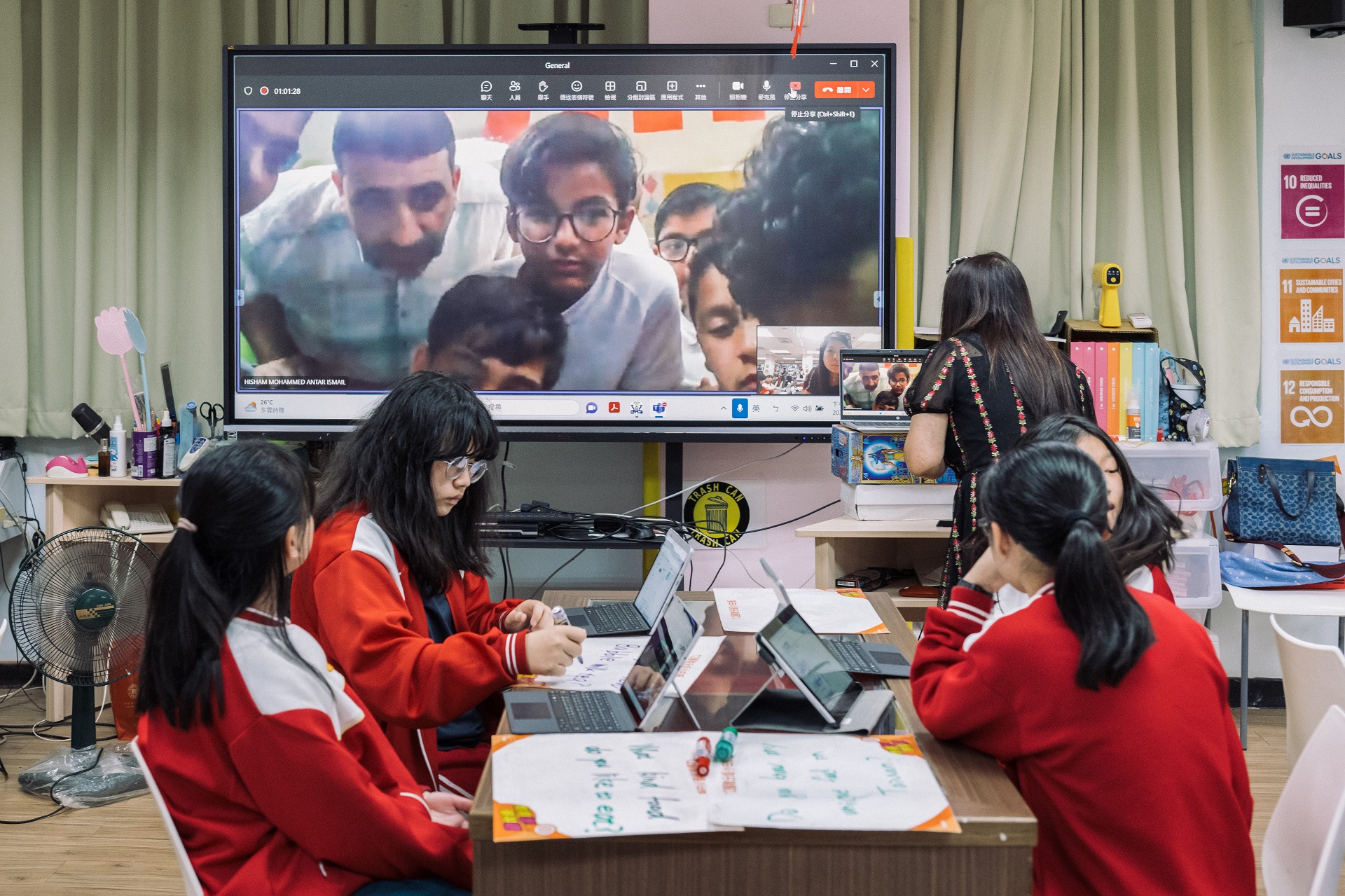 Students in a classroom interact virtually with other students visible on a screen.