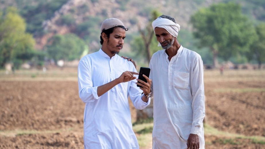 Two farmers standing in an arid village field looking at a mobile phone.