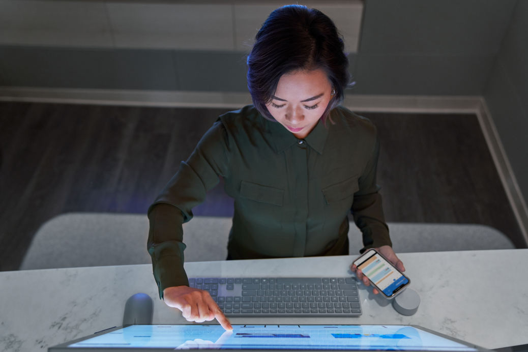 Top down view of a woman wearing a dark shirt in a dim office scrolling or working on a Microsoft Surface Studio and holding a phone