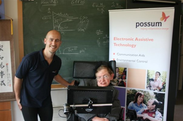 Two men, Hector Minto and late Professor Stephen Hawking, smiling at the camera. Also seen is Hawking's communication aid. 