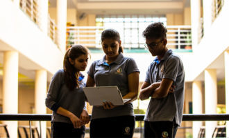 A group of students reviews notes on a laptop together while standing in hallway of private school in India.