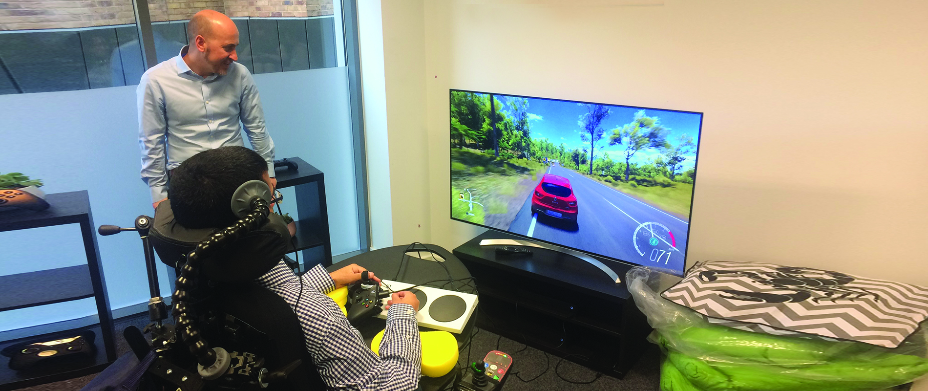 Hector Minto looks on as a gamer with disabilities uses XBox adaptive controller. The device screen displays a car racing game.