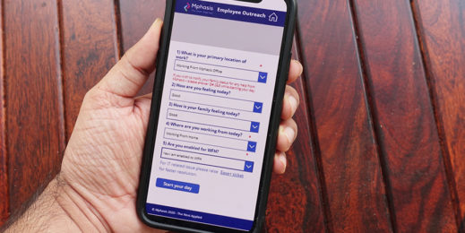 An employee using the 'Employee Outreach' app of Emphasis. On the device screen, the app shows various questions, like 'What is your primary location of work?', 'How are you feeling today?', 'How is your family feeling today?', and 'Are you enabled for WFH?'. Each question has a dropdown menu, allowing the users to choose relevant answers. The bottom corner of the app shows the final step - 'Start your day'.