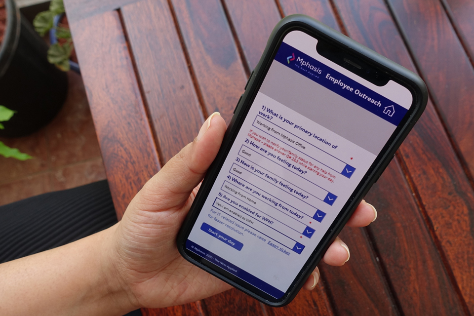 An employee using the 'Employee Outreach' app of Emphasis. On the device screen, the app shows various questions, like 'What is your primary location of work?', 'How are you feeling today?', 'How is your family feeling today?', and 'Are you enabled for WFH?'. Each question has a dropdown menu, allowing the users to choose relevant answers. The bottom corner of the app shows the final step - 'Start your day'.