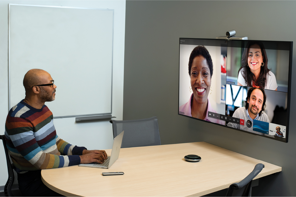 photo of a man sitting in a meeting room attending a Microsoft Teams call with other participants visible on a display