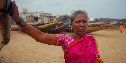 an old woman wearing a bright pink sari at the beach with fishing boats in the backdrop