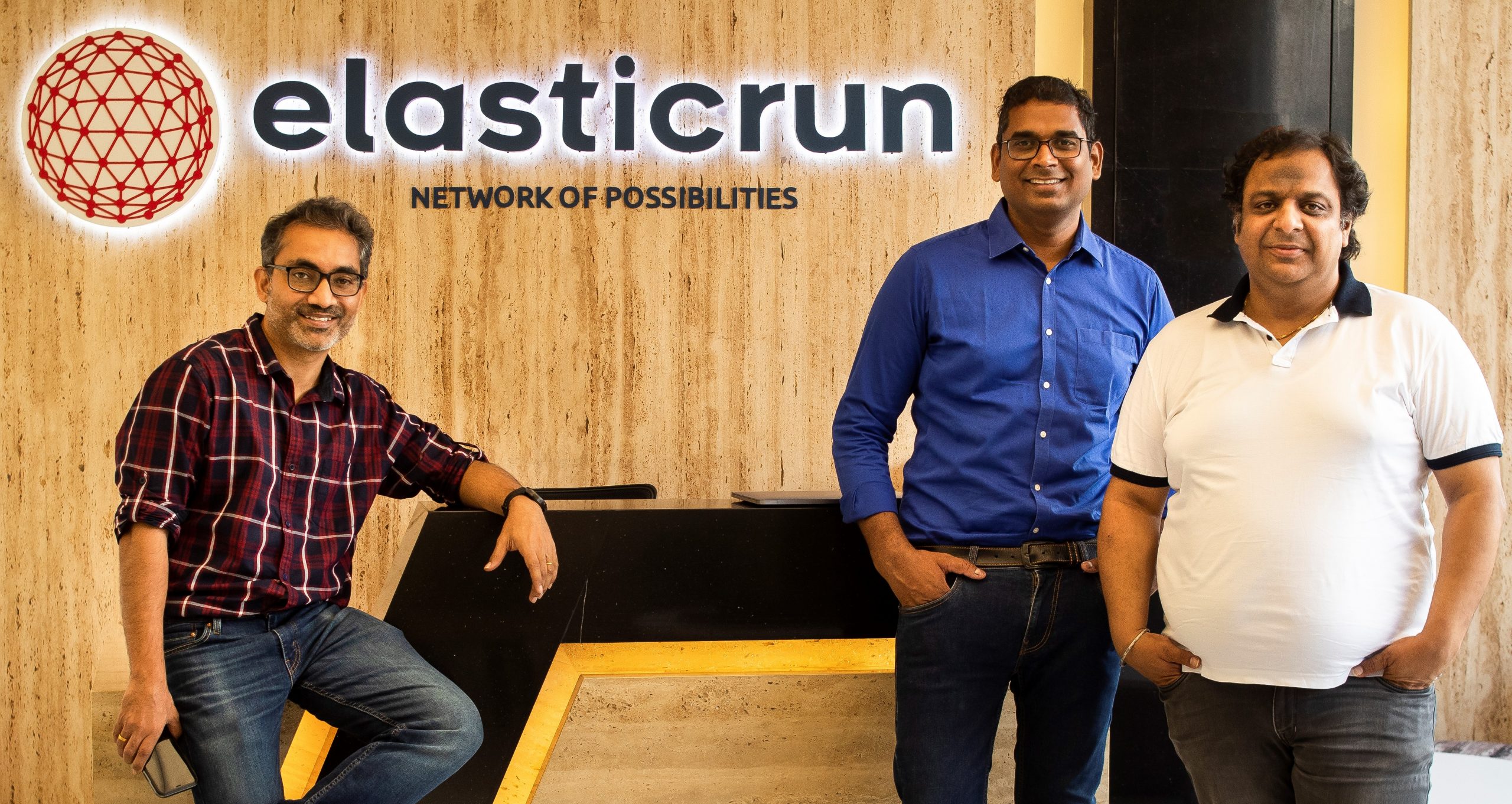Three men standing in front of a wall with ElasticRun's logo