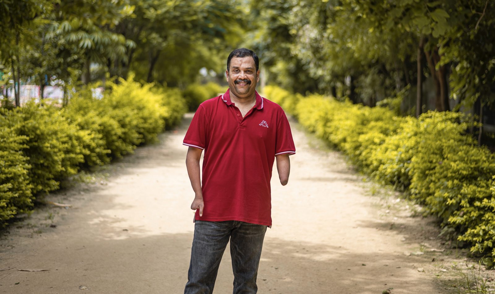 a man wearing a red t-shirt standing on a pathway lined with hedges and trees