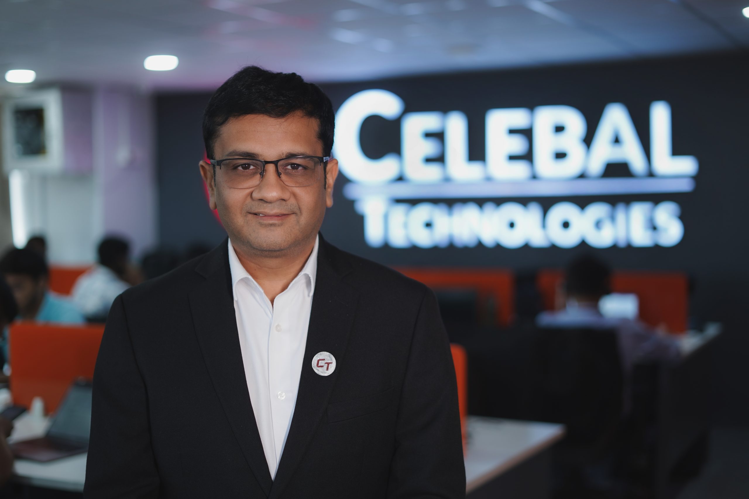 a man wearing a black jacket, white shirt, and glasses looking at the camera with Celebal Technologies signboard in the background