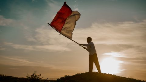 A photo of a student waving the Indonesian flag on a hill at sunset