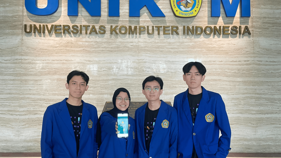 Four students in blue blazers, one of them showing a mobile app interface, standing in front of the university's logo on a wall.