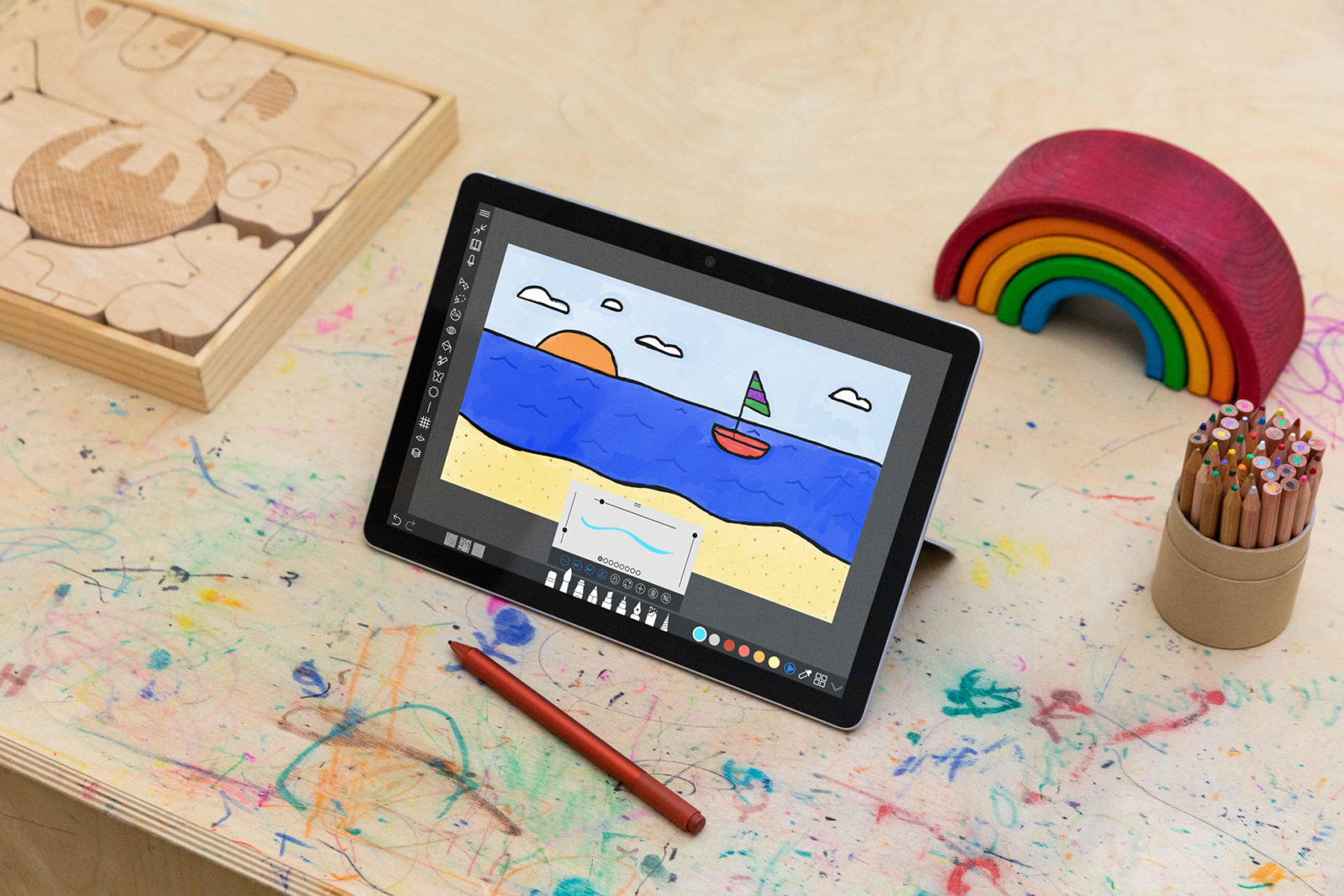 На детском столе карандаши, логические игрушки и планшет на базе Windows 10. На экране – рисунок в программе paint