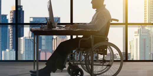 Мужчина в инвалидном кресле работает за компьтеным столом на фоне стеклянного окна, за которым виден большой город