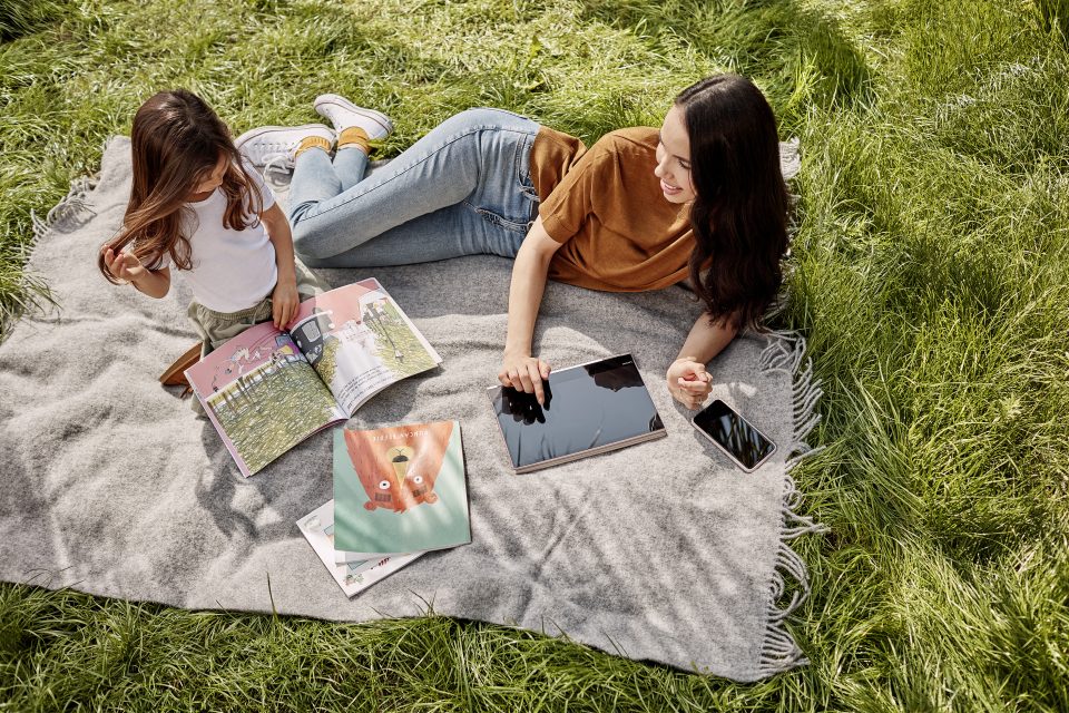 Žena leží na dece na trávě se svou dcerou. Žena ukazuje na tablet, dcera si prohlíží časopis.