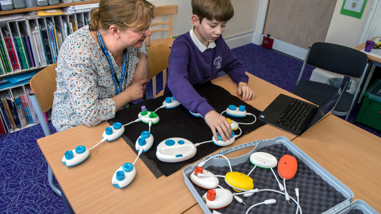 Theo sits at a desk with his teacher Allison with his hands on a Code Jumper pod