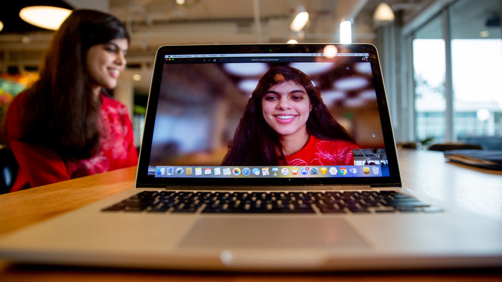 A laptop showing a screenshot of Swetha Machanavajhalar on Skype with background blur, Swetha is live in the background behind the laptop being recorded on an unseen mobile device