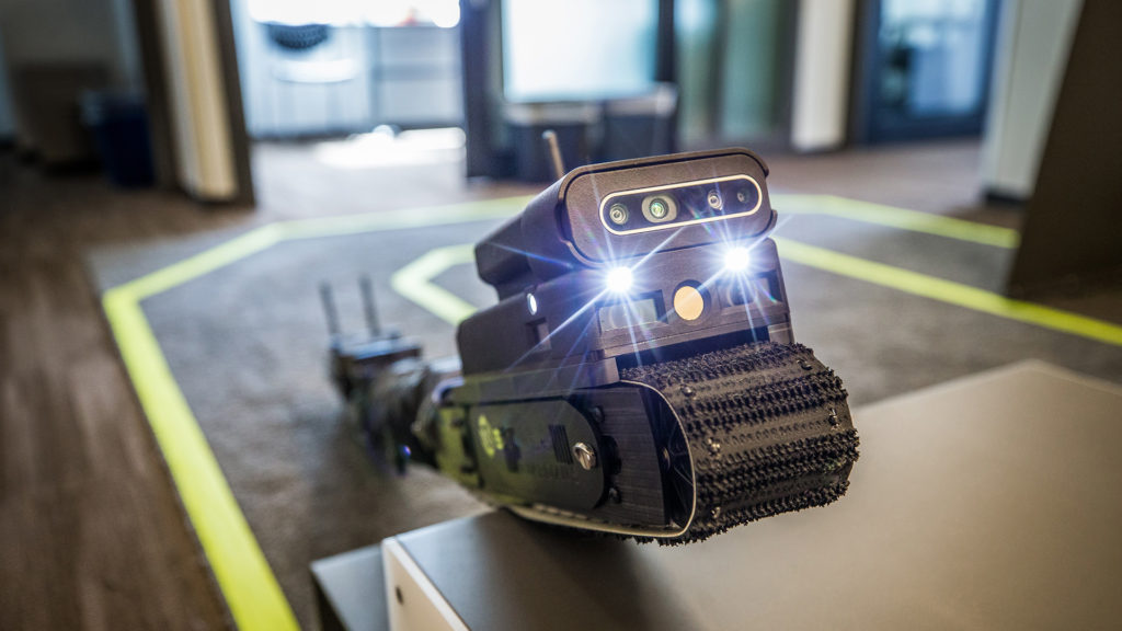 A Sarcos Guardian S robot climbs on a object in a test environment, two of its front lights shining