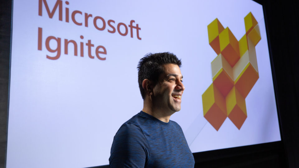 Rohan Kumar, corporate vice president for Azure Data, stands in front of a screen with the words “Microsoft Ignite”