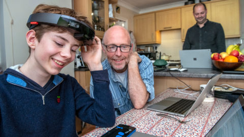 Theo smiles with his left hand touching a HoloLens that sits on his head, while interacting with Microsoft researchers at a kitchen table