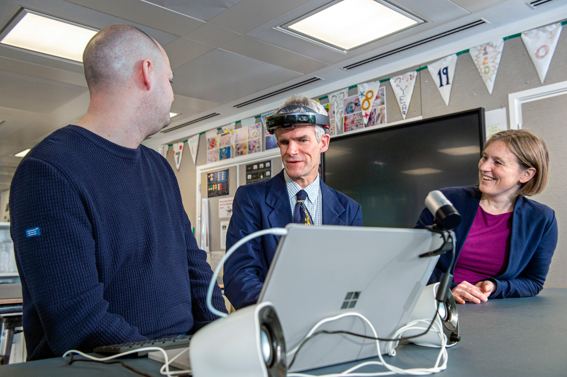 Peter Bosher interacts with Microsoft researchers while wearing a HoloLens, sitting at a table in a research lab, with a Microsoft Surface Book computer in the foreground