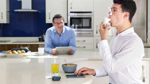 A man watching a teenage boy use a spirometer in a kitchen