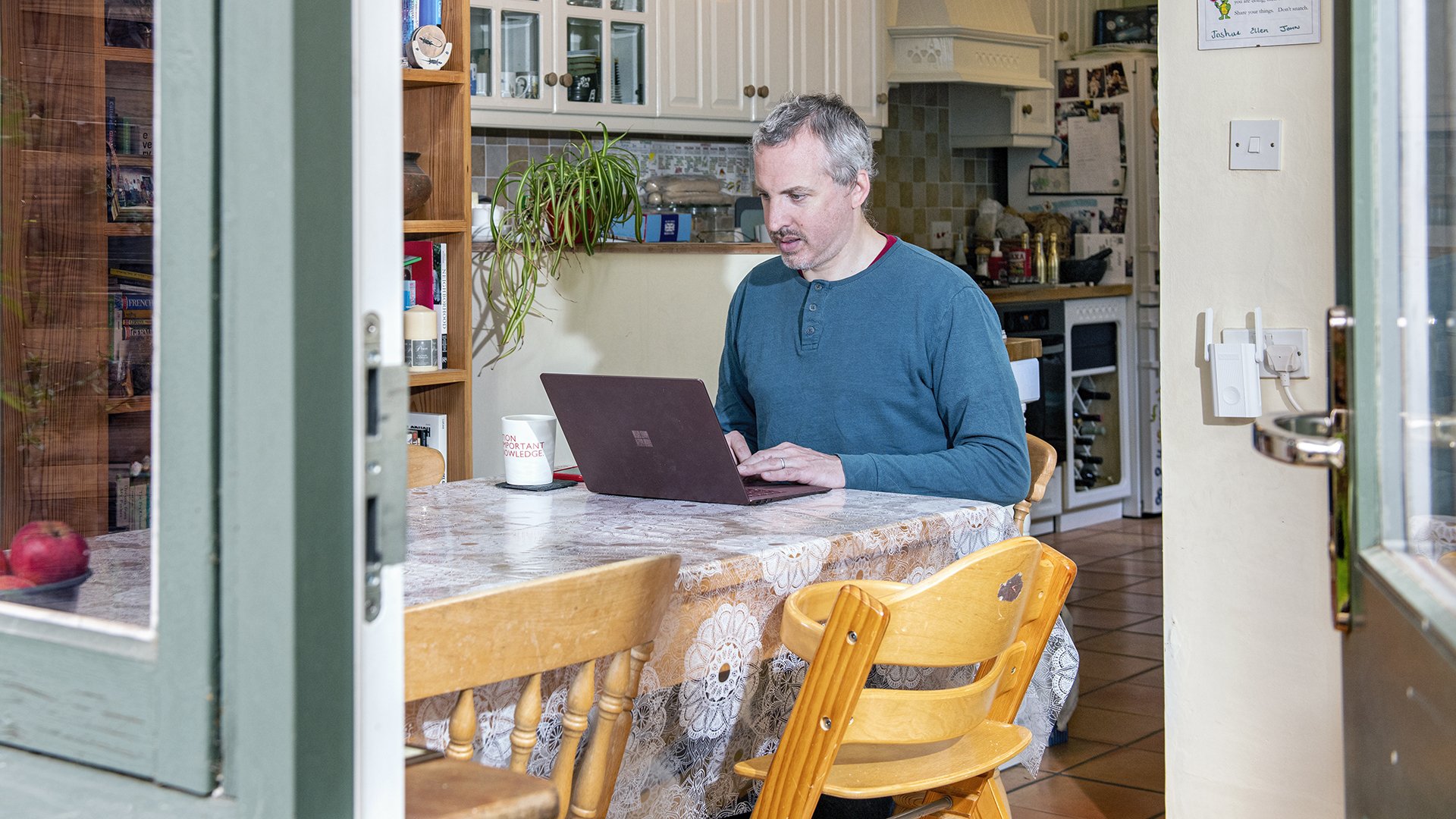 A man sits at a kitchen table working on a laptop