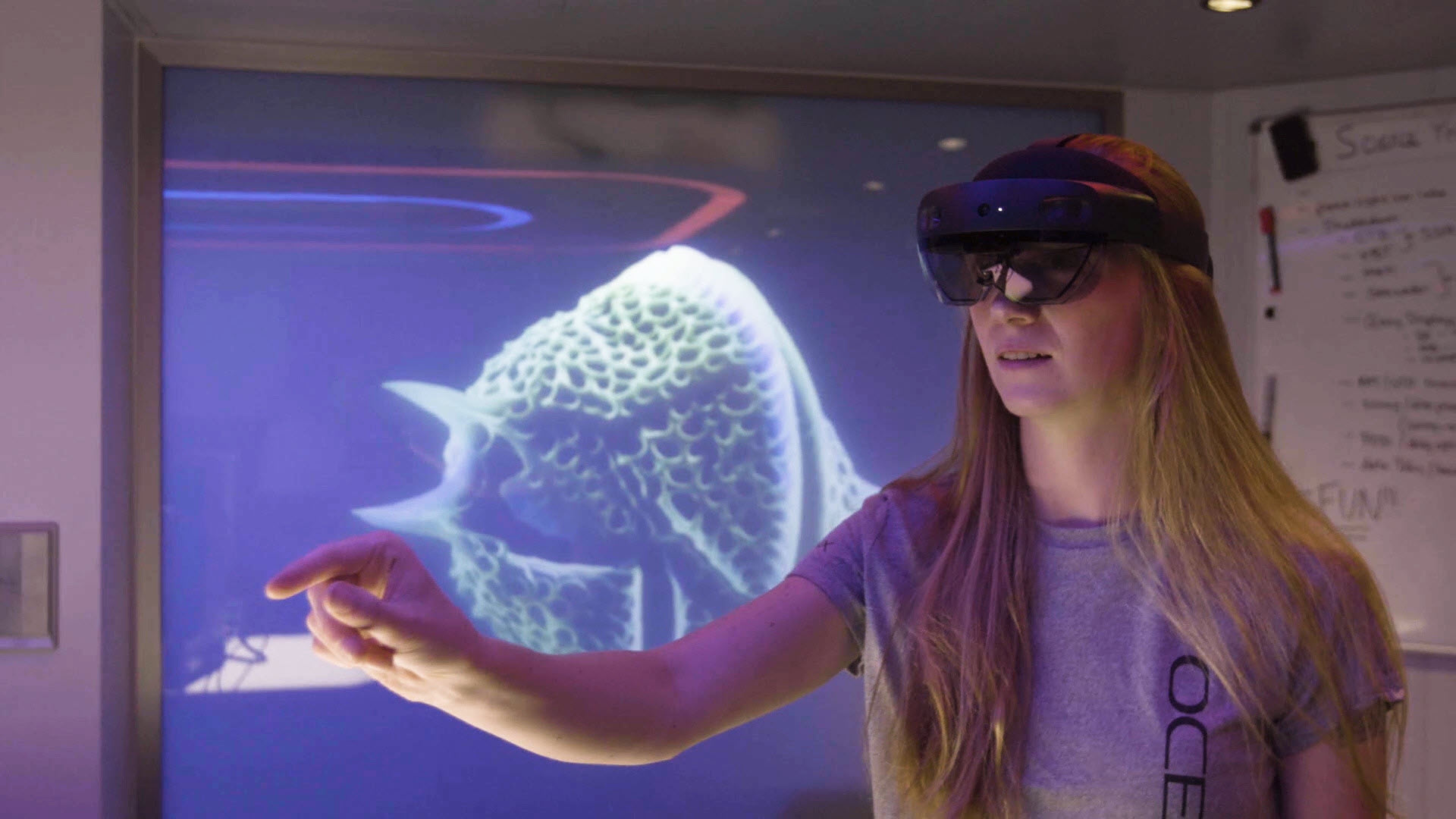 A woman wearing an OceanX shirt and a HoloLens interacts with an unseen hologram with an image of sea life in the background.
