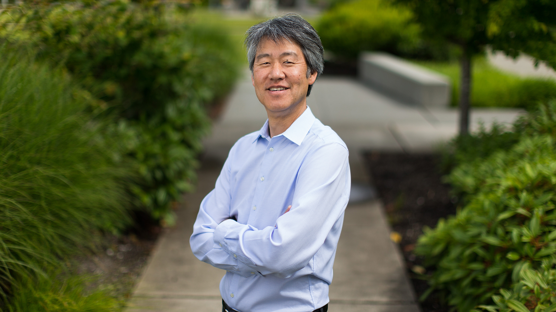 Peter Lee stands with arms crossed on a sidewalk surrounded by greenery