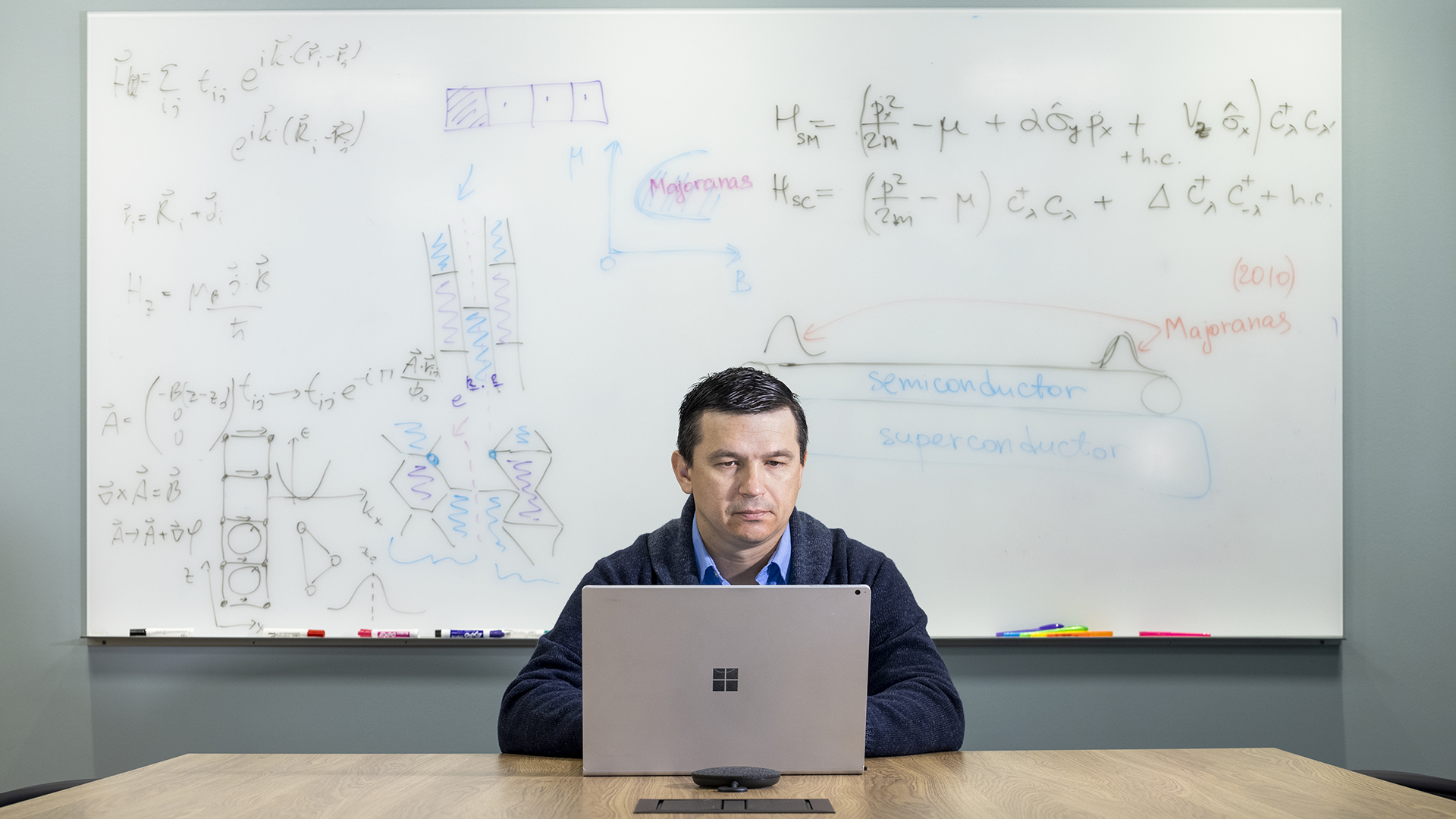 A man sits at a desk on a computer with a whiteboard behind him