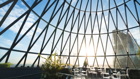 The Gherkin building. restaurant tables at bottom and photo is filled with a giant glass wall and ceiling, with giant metal criss cross holding it up, and sun and clouds filling up the sky behind it.