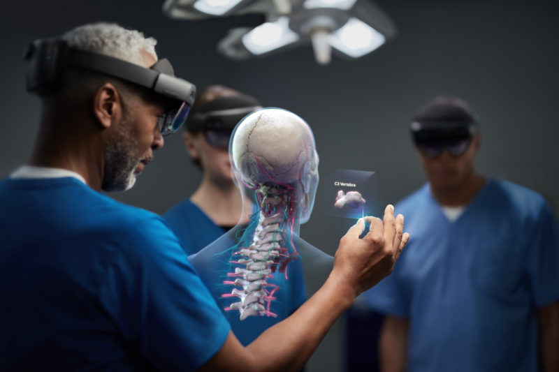 A doctor wearing HoloLens 2, with two other doctors in the background, view a hologram of a human spine