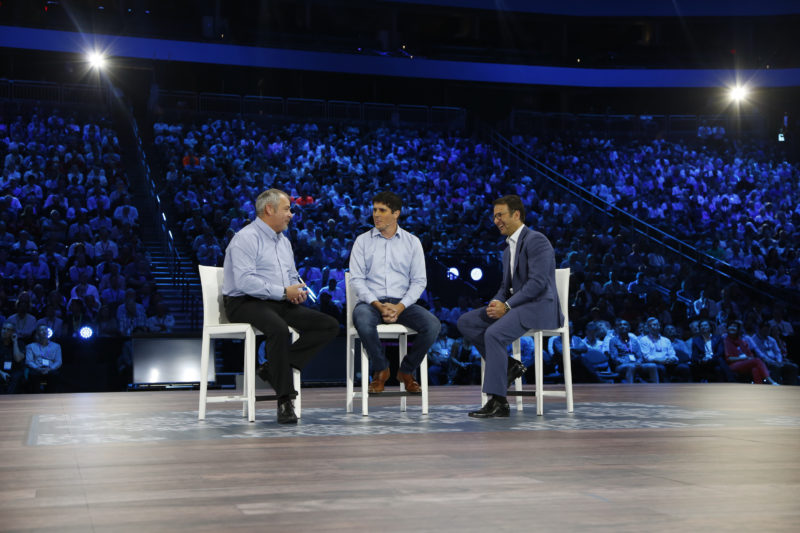 Three men sit on chairs on stage speaking.
