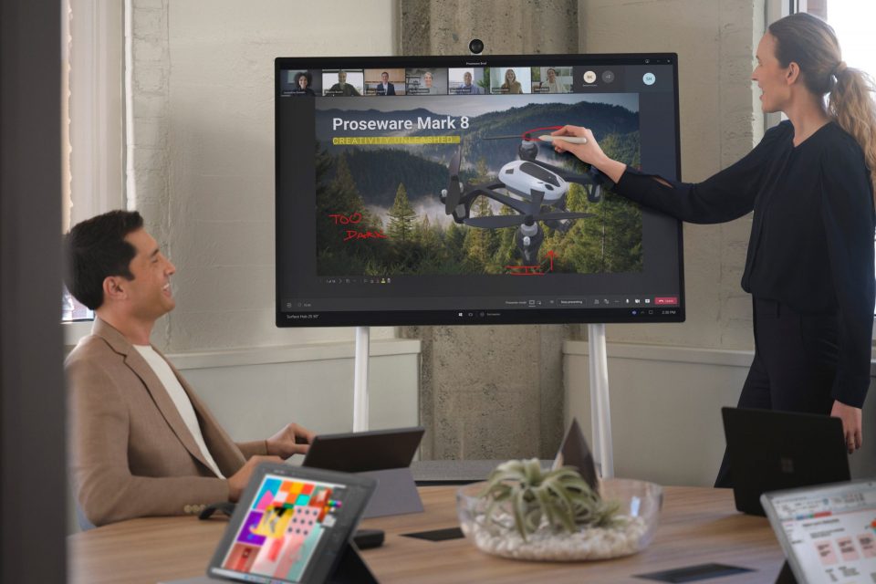 A presenter uses a Surface Hub 2 Pen to take notes in Whiteboard while attending a Teams meeting with remote and in-person colleagues.