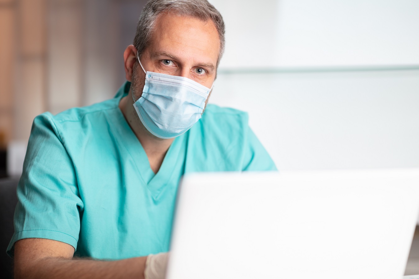 Lekarz w stroju medycznym i maseczce ochronnej korzystający z laptopa w swoim gabinecie