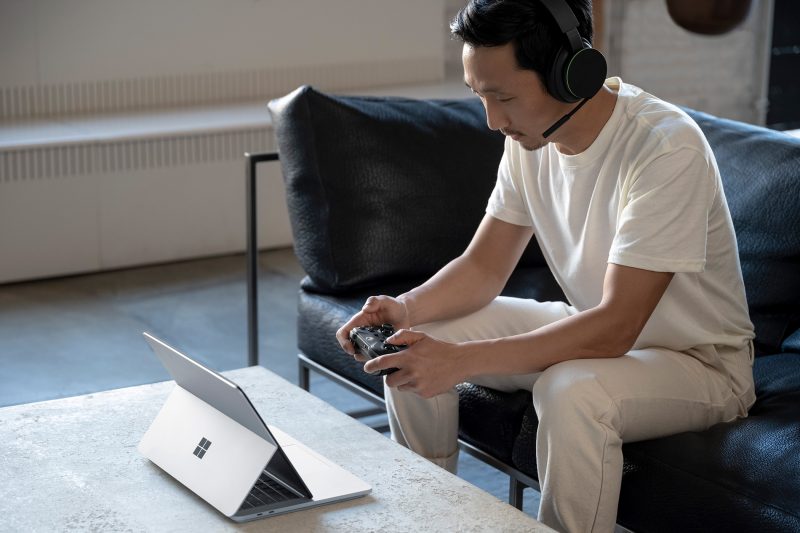 A man sitting down using an Xbox controller with a headset on while on a Surface laptop