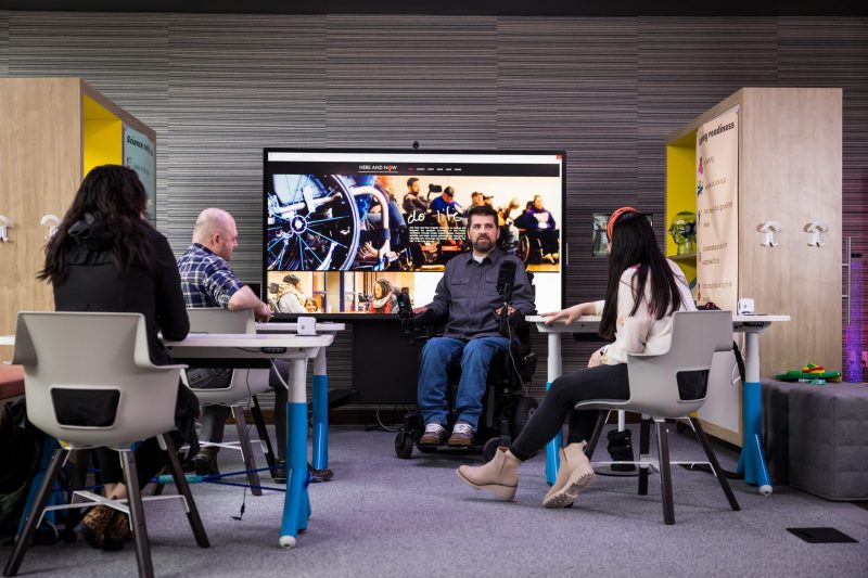 Four people sitting inside the Inclusive Tech Lab at desks; having a conversation and looking at a large screen