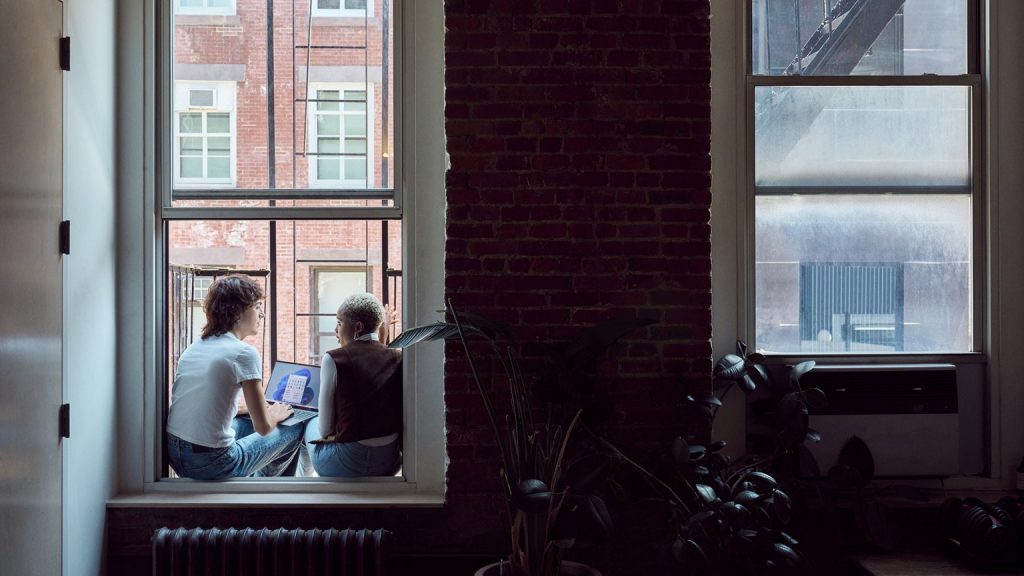 Two women sit on a window sill talking, one with a laptop in her lap