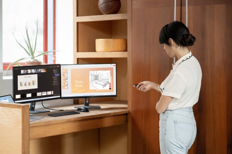 A person stands in front of two computer monitors holding a device while on a video call.