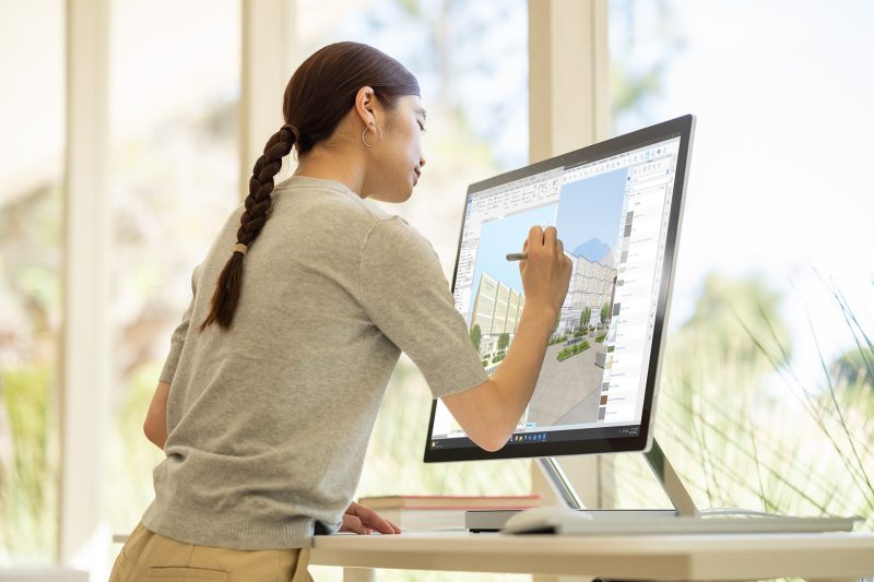 A person is drawing out the design of a building on the display of an all-in-one computer while standing over a desk.