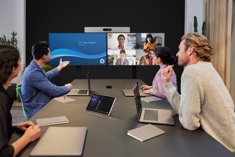 Four people sit at a table with laptops and a tablet, as they interact with a monitor that shows four people on the screen.